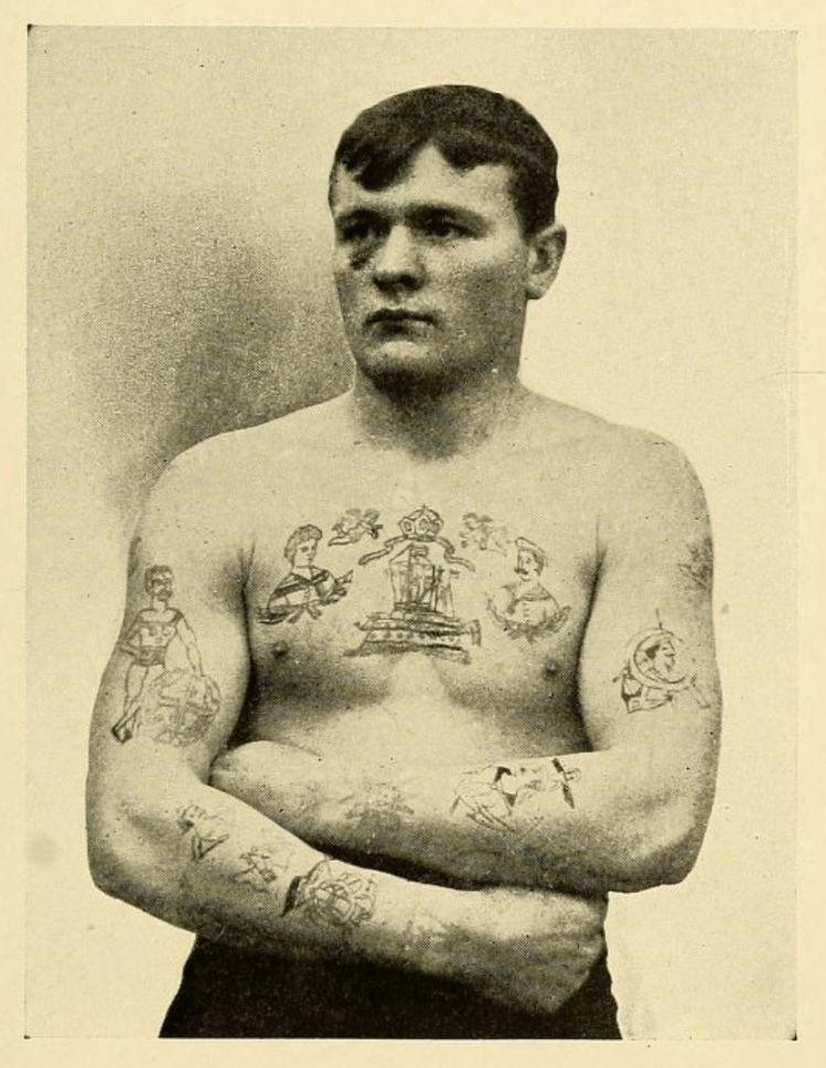 a photograph of a white man with a black eye and arms folded. On his chest and arms are several naval tattoos